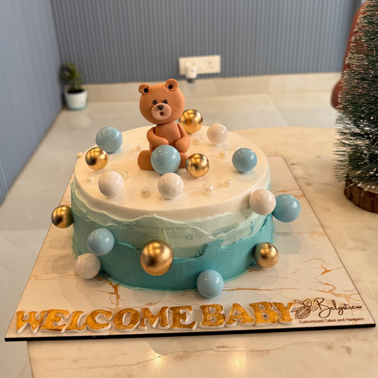 A bespoke Belgstrew Welcome Baby Boy cake featuring a handcrafted blue teddy bear topper on a soft pastel-colored sponge.