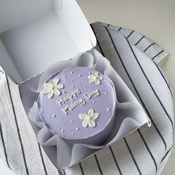 A soft lilac bento cake with white piped daisies, white pearls, and "Happy Mommy Day" written in white icing inside a white box.