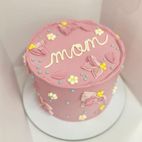 A tall mauve cake with textured palette-knife floral petals, small white piped daisies, teal accents, and "mom" written in white cursive.