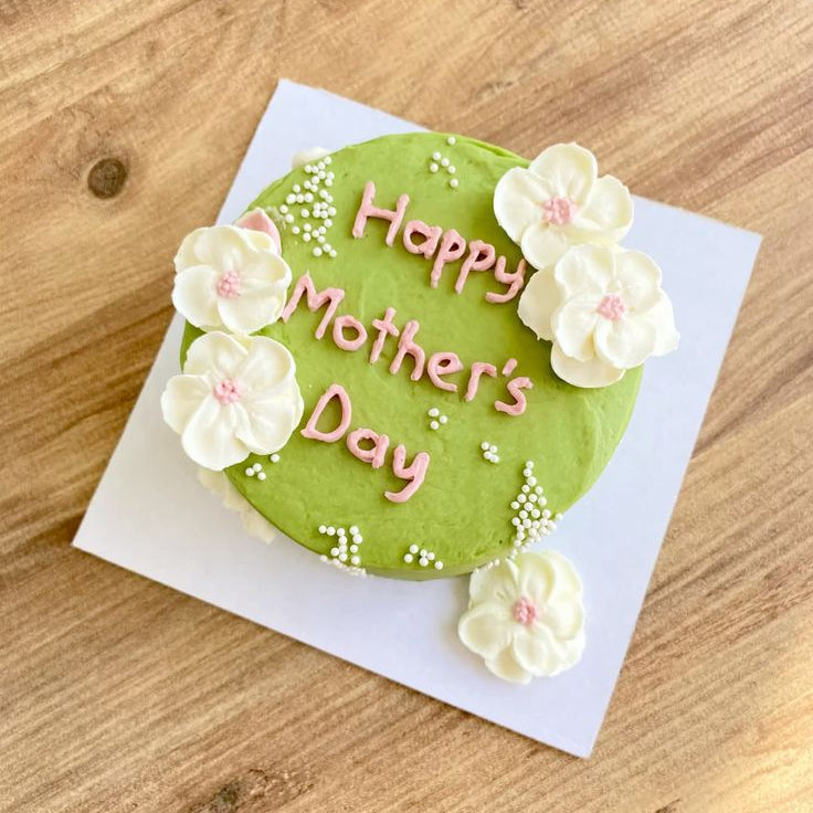 A vibrant green cake decorated with large white piped flowers, small white pearls, and "Happy Mother's Day" in pink icing.