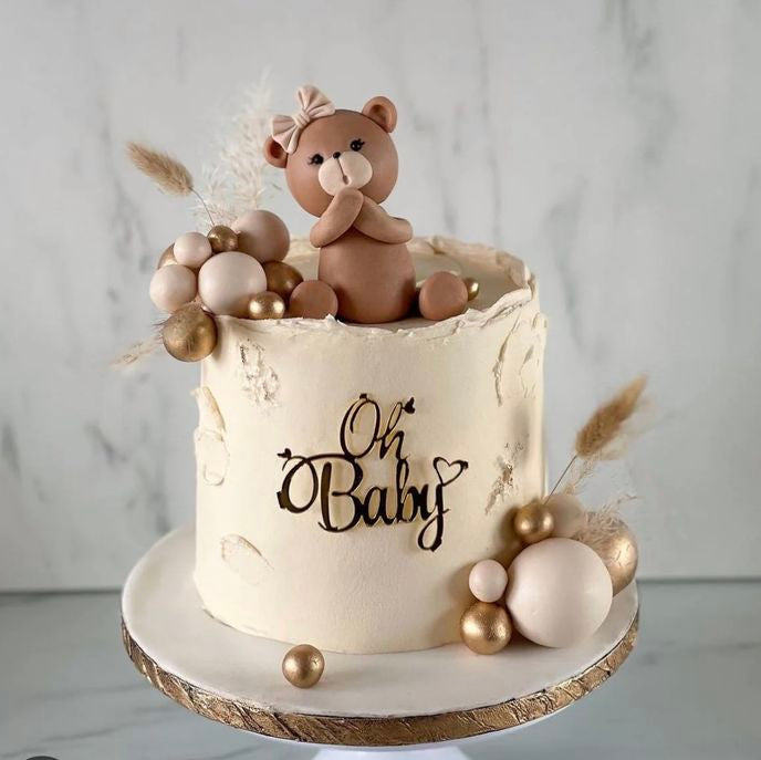 A tall rustic ivory baby shower cake decorated with a mirror gold Oh Baby plaque, cream and gold chocolate spheres, dried pampas grass, and a hand-made teddy bear wearing a bow on top.