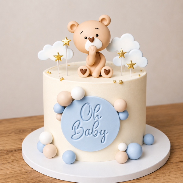 A smooth ivory baby shower cake with a blue 'Oh Baby' plaque, decorated with blue and beige chocolate spheres, topped with a fondant teddy bear, clouds, and gold stars.