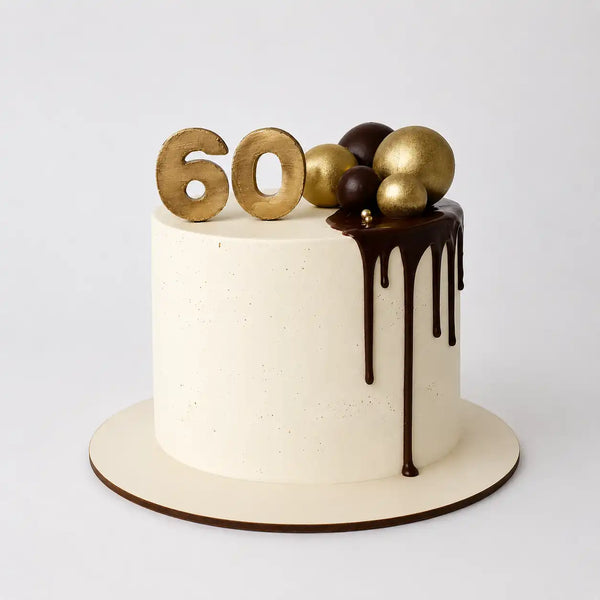 A tall round 60th birthday cake for him with a smooth white cream base, featuring a dark chocolate drip down the side, topped with large gold and brown spheres and a gold number 60.