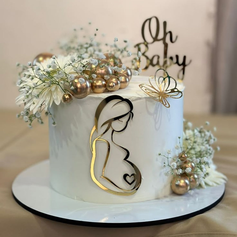 A premium white baby shower cake decorated with a mirror gold silhouette of a pregnant woman, fresh white flowers, baby's breath, metallic gold spheres, and an Oh Baby topper.