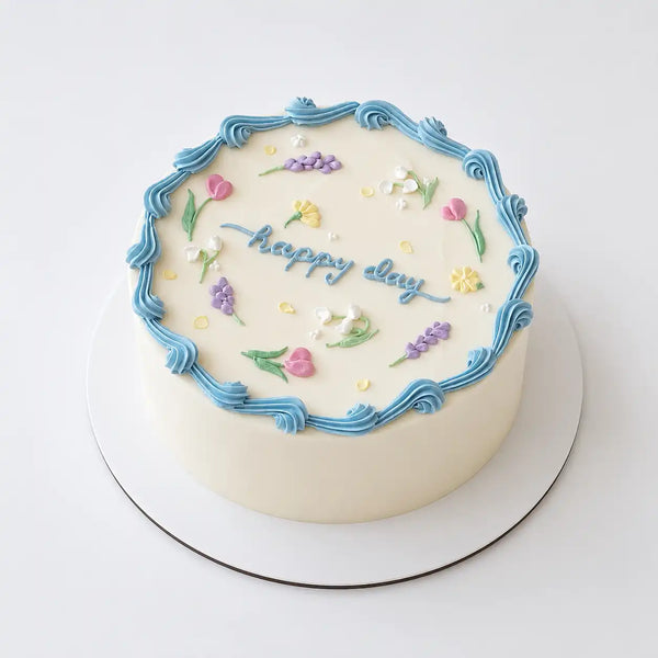 A minimalist white round Korean style cake decorated with a wavy blue buttercream border, small hand-piped pastel flowers like tulips and lavender, and blue cursive writing reading happy day.