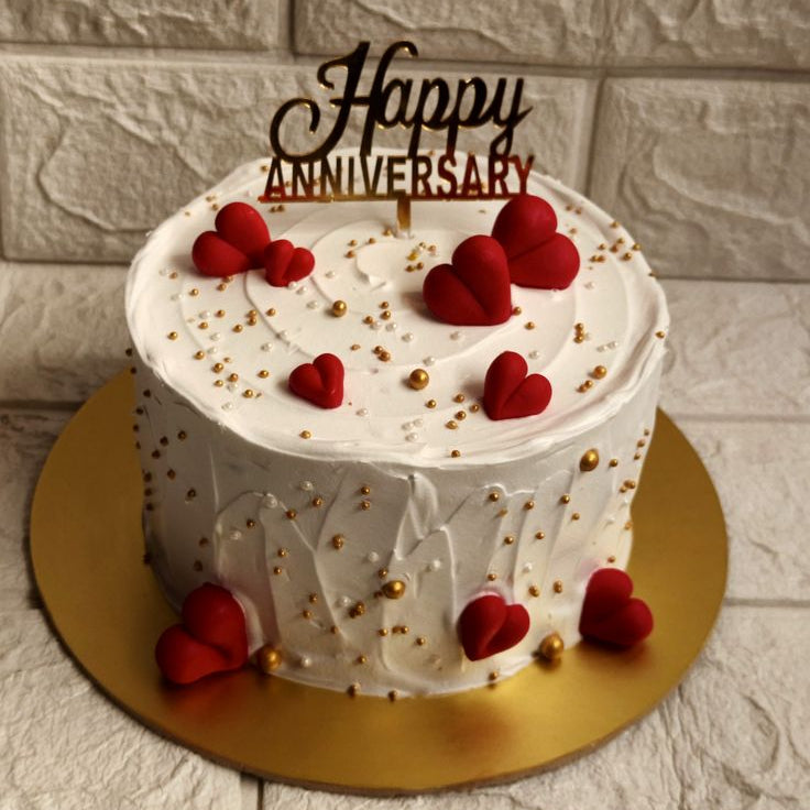 A white frosted anniversary cake with a textured swirl design, decorated with scattered 3D red fondant hearts, gold sprinkles, and a shiny gold Happy Anniversary topper.