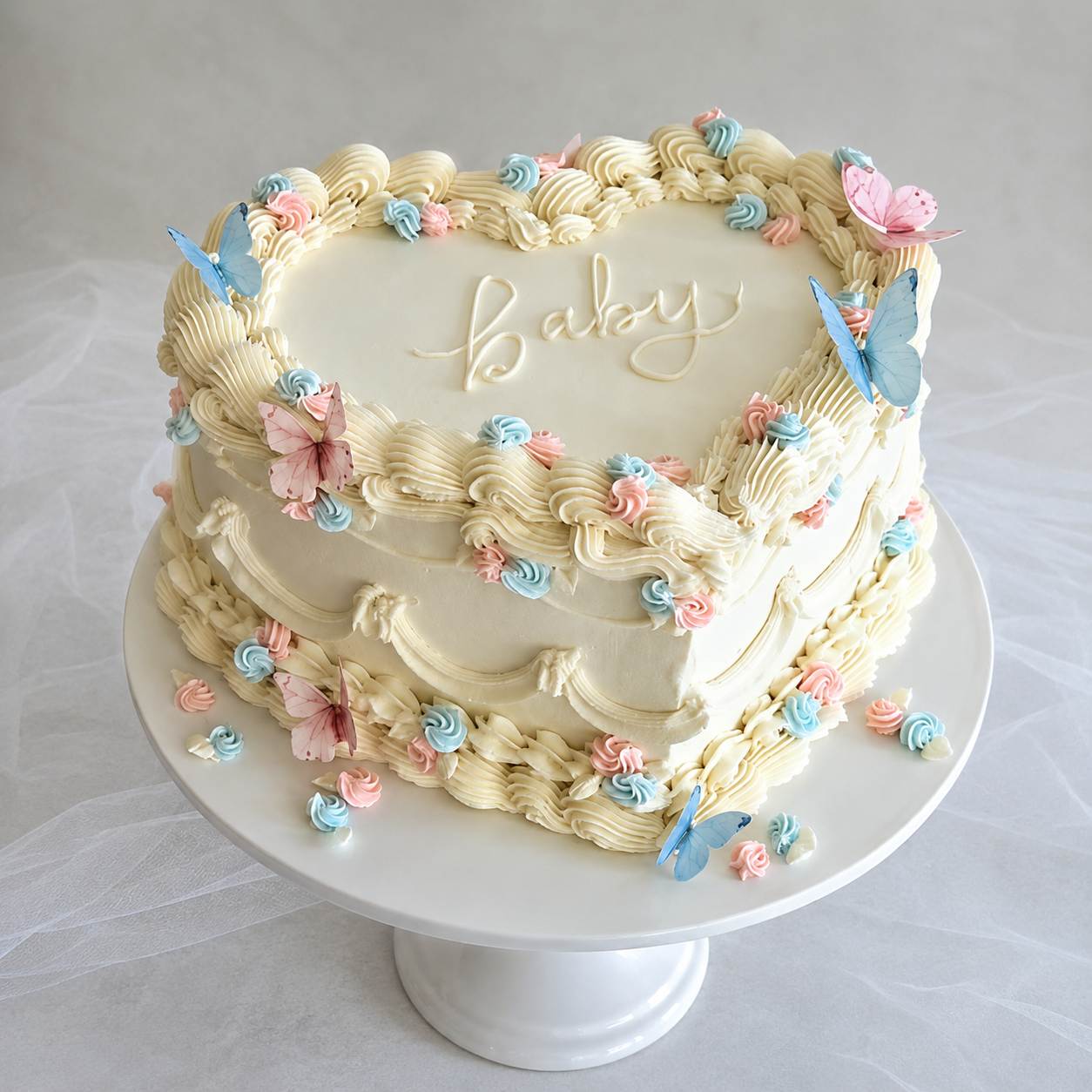 An elegant heart-shaped ivory vintage cake with detailed ruffle piping, decorated with pastel pink and blue butterflies and the word baby written in cursive.