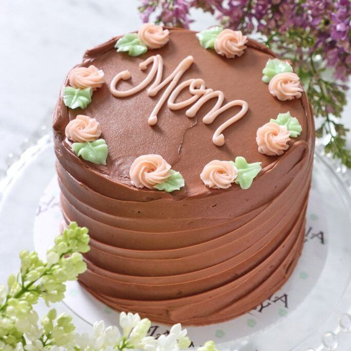 A 1kg chocolate cake with horizontal pleated frosting, peach-colored piped roses, and "Mom" written in pink icing.