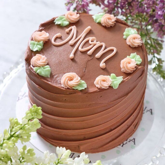A 1kg chocolate cake with horizontal pleated frosting, peach-colored piped roses, and "Mom" written in pink icing.