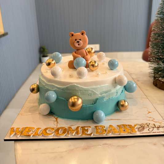 A bespoke Belgstrew Welcome Baby Boy cake featuring a handcrafted blue teddy bear topper on a soft pastel-colored sponge.