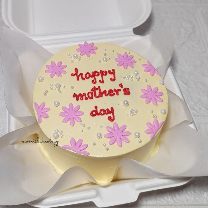 A cream-colored bento cake with pink hand-piped daisies, white pearls, and "happy mother's day" written in red icing.