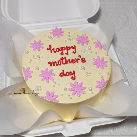 A cream-colored bento cake with pink hand-piped daisies, white pearls, and "happy mother's day" written in red icing.