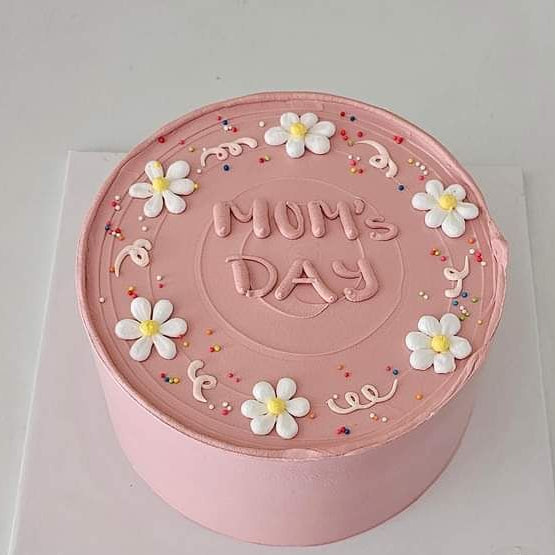 A dusty pink monochromatic cake with hand-piped white daisies, "Mom's Day" written in pink icing, and colorful sprinkles.