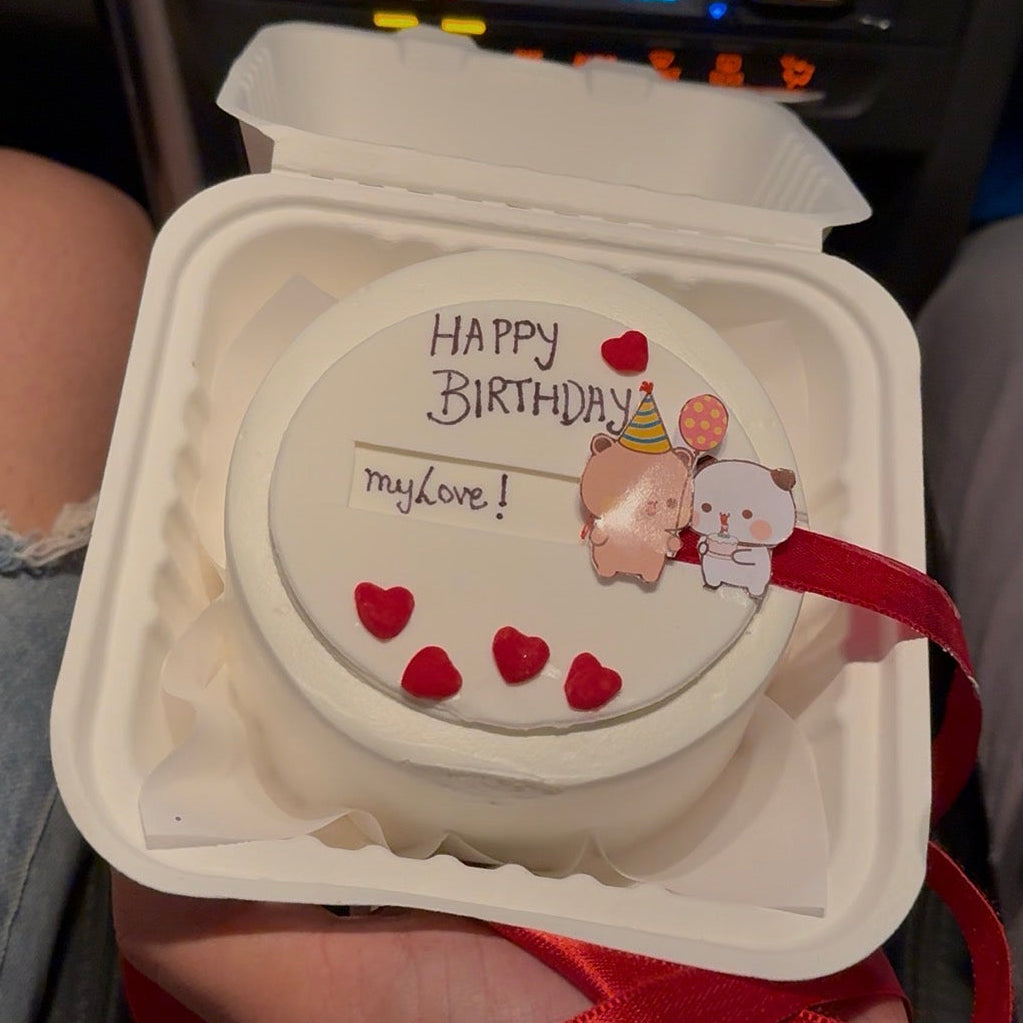 A handcrafted 100% eggless Bubu Dudu bento cake featuring a white frosting base, hand-painted Bubu and Dudu bear characters, and a satin ribbon pull mechanism for a surprise message reveal.