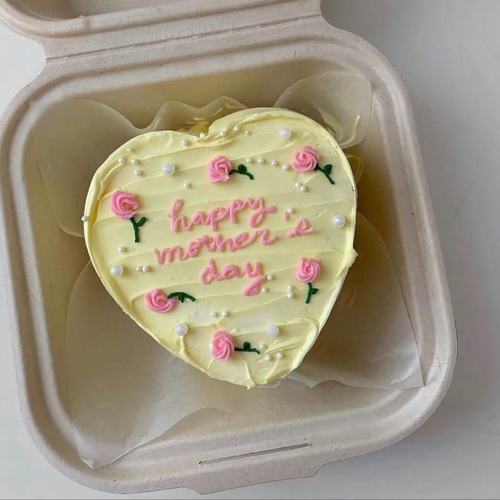 A heart-shaped yellow bento cake with pink piped rosebuds, white pearls, and "happy mother's day" written in pink script.