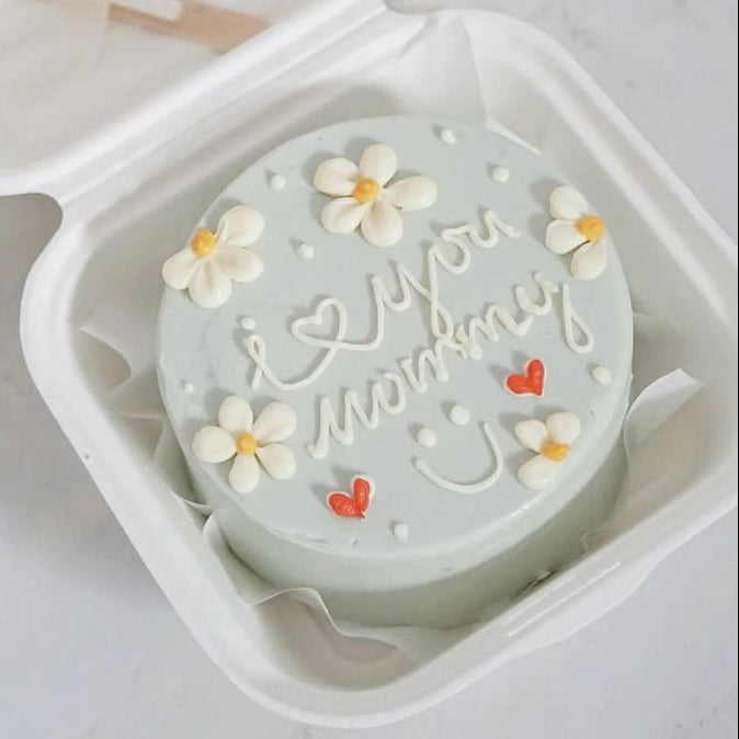 A muted sage green bento cake with white piped daisies, red hearts, and "i ♡ you mommy" script with a smiley face.