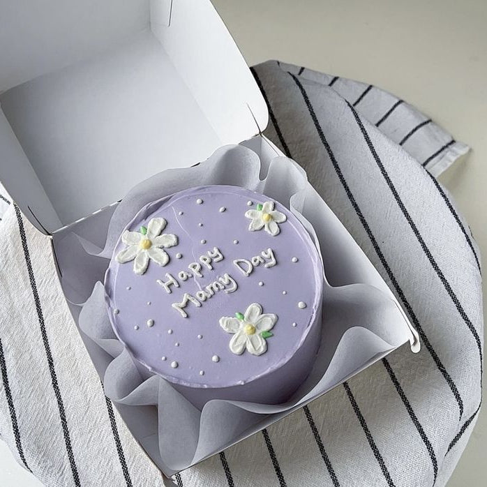 A soft lilac bento cake with white piped daisies, white pearls, and "Happy Mommy Day" written in white icing inside a white box.