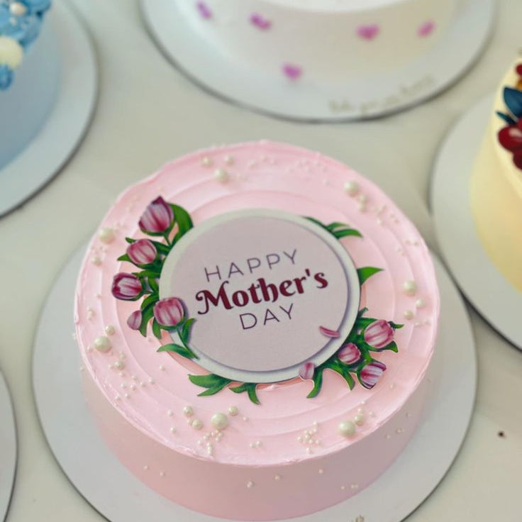 A soft pink textured cake decorated with white edible pearls and a "Happy Mother's Day" floral plaque.