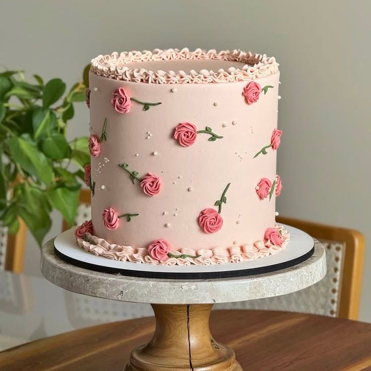 A tall blush-pink cake decorated with hand-piped pink roses, green stems, white pearls, and ruffled shell-piped borders.
