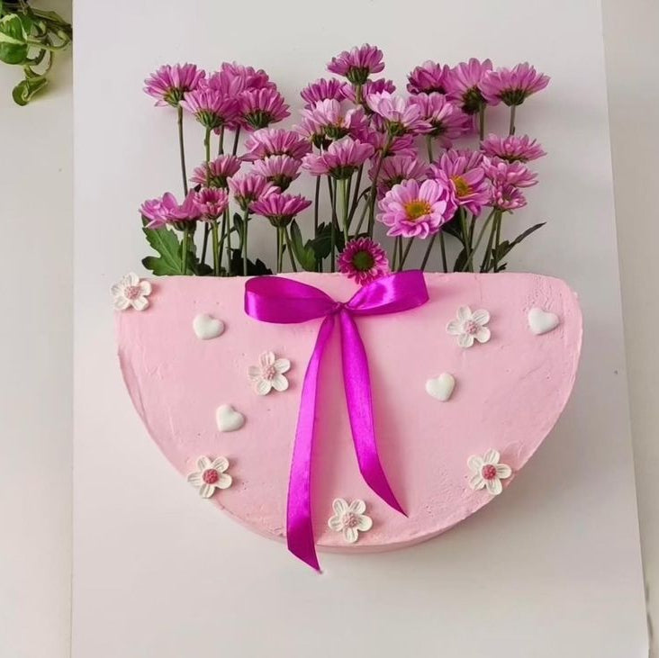 A vertical pink half-circle cake designed as a flower bag, overflowing with fresh purple chrysanthemums and decorated with a magenta ribbon.