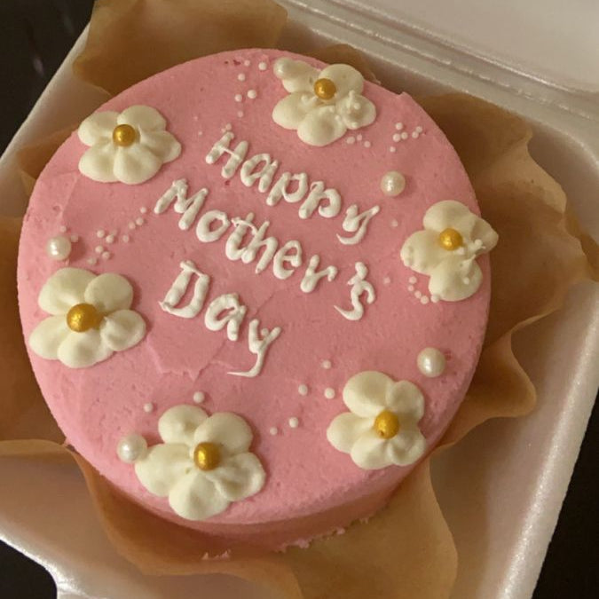 A vibrant pink bento cake with white piped flowers, gold centers, and white pearls featuring "Happy Mother's Day" script.