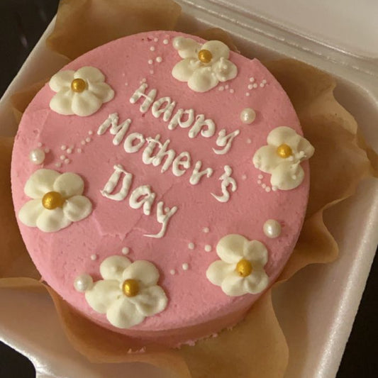 A vibrant pink bento cake with white piped flowers, gold centers, and white pearls featuring "Happy Mother's Day" script.