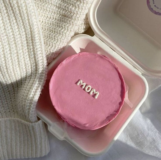 A vibrant pink textured bento cake in a white box with "mom" hand-piped in white lowercase letters.