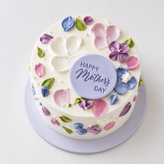 A white cake decorated with thick, textured frosting strokes in lavender, blue, and pink to look like flower petals, with a central "Happy Mother's Day" plaque.