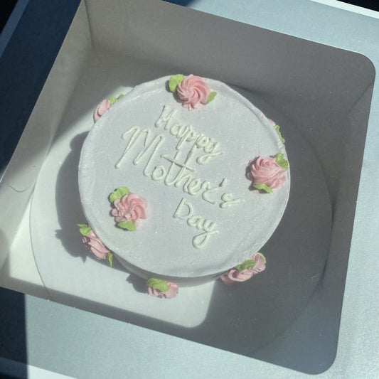 A white cake with a shimmer finish, decorated with pink piped rosettes and "Happy Mother's Day" written in white icing.
