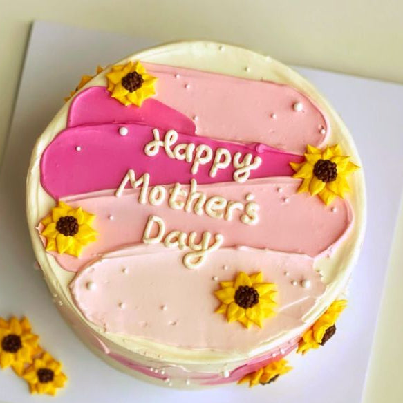 A white cake with artistic pink and magenta frosting strokes, decorated with yellow piped sunflowers and a Happy Mother's Day message.