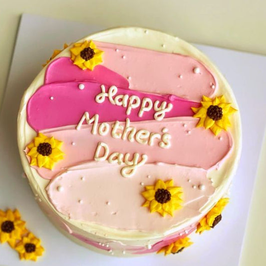 A white cake with artistic pink and magenta frosting strokes, decorated with yellow piped sunflowers and a Happy Mother's Day message.