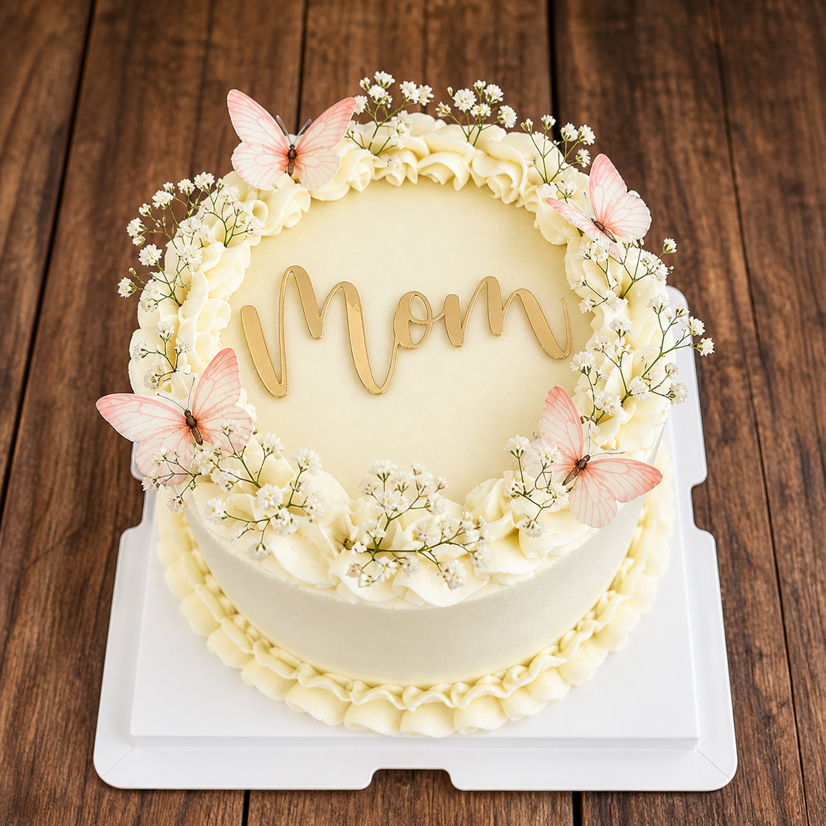 A white ruffled cake decorated with fresh baby's breath flowers, peach butterflies, and a gold 'Mom' topper on a wooden background.