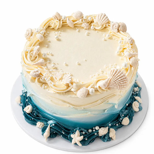 A tall premium round ocean themed cake with an ivory top transitioning into turquoise and dark teal waves, decorated with piped ruffles, white pearls, and hand-molded chocolate seashells as seen in image.