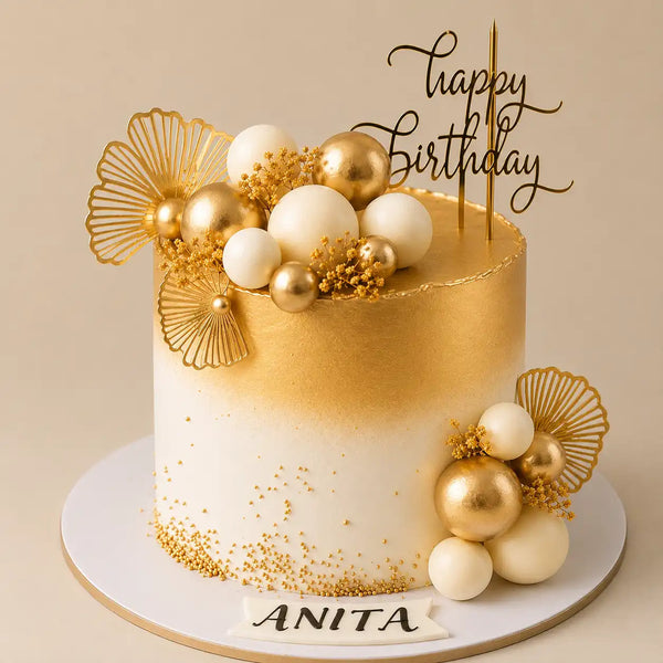 A tall round premium birthday cake with a smooth white bottom half transitioning to a bright painted gold top half, decorated with white and polished gold chocolate spheres, delicate yellow baby's breath, metallic gold fan leaves, and a name banner reading Anita as seen in image.