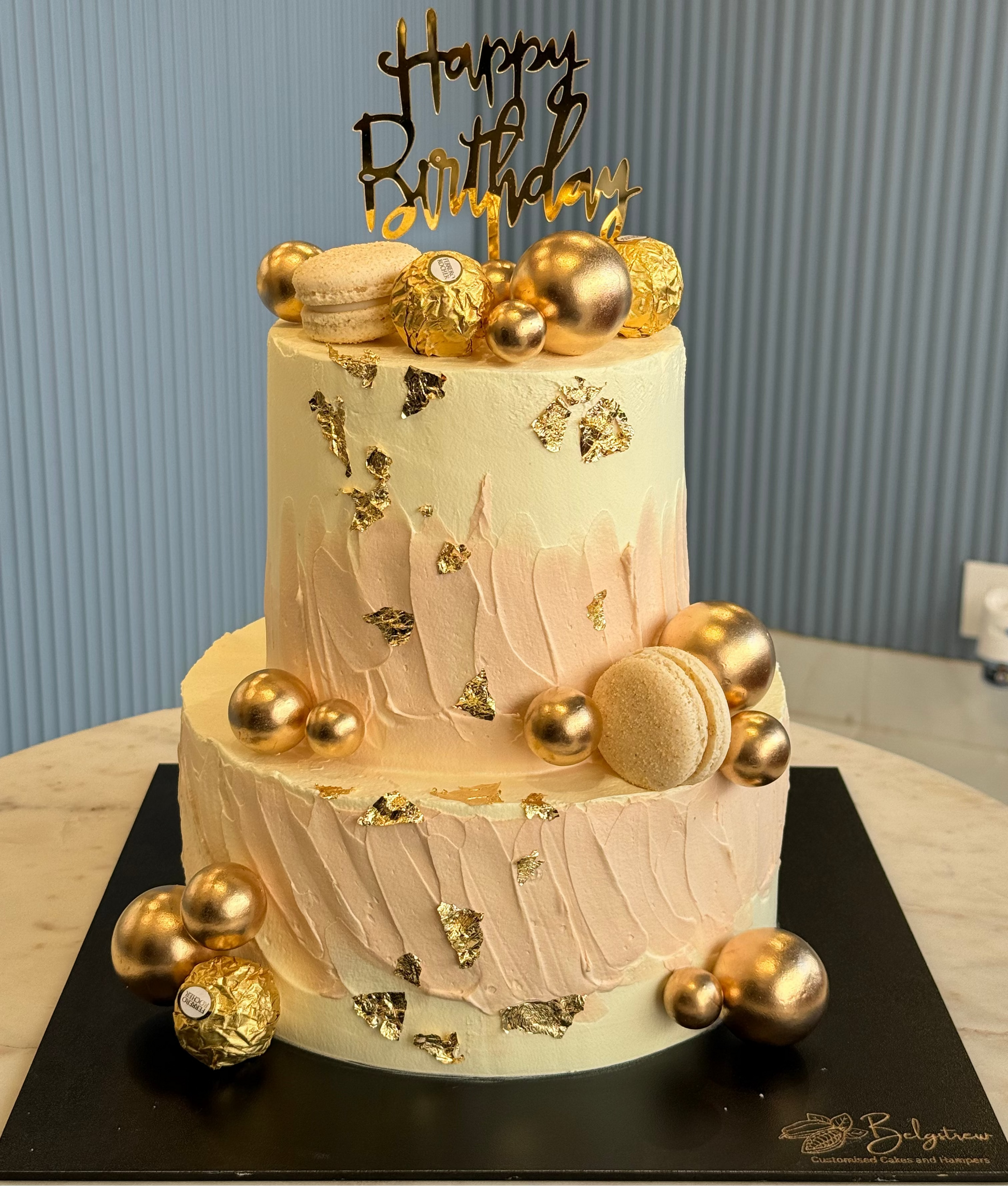 Two-tier birthday cake with gold decorations and 'Happy Birthday' topper on a striped background