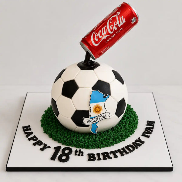 A fully sculpted 3D black and white spherical football cake on a bed of green grass frosting, featuring a custom Argentina flag map on the front and a gravity-defying red Coca-Cola can floating in mid-air above it as seen in image.
