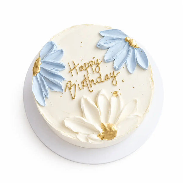 A minimalist round white birthday cake with three 3D palette-knife sculpted flowers in light blue and white, decorated with hand-painted gold centers and gold cursive text reading Happy Birthday as seen in image.