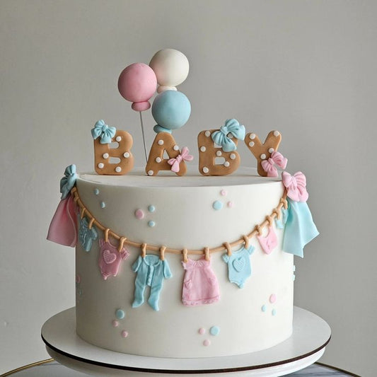 A smooth ivory baby shower cake decorated with a miniature clothesline hanging pink and blue baby clothes, topped with custom BABY cookie letters and three pastel balloons.