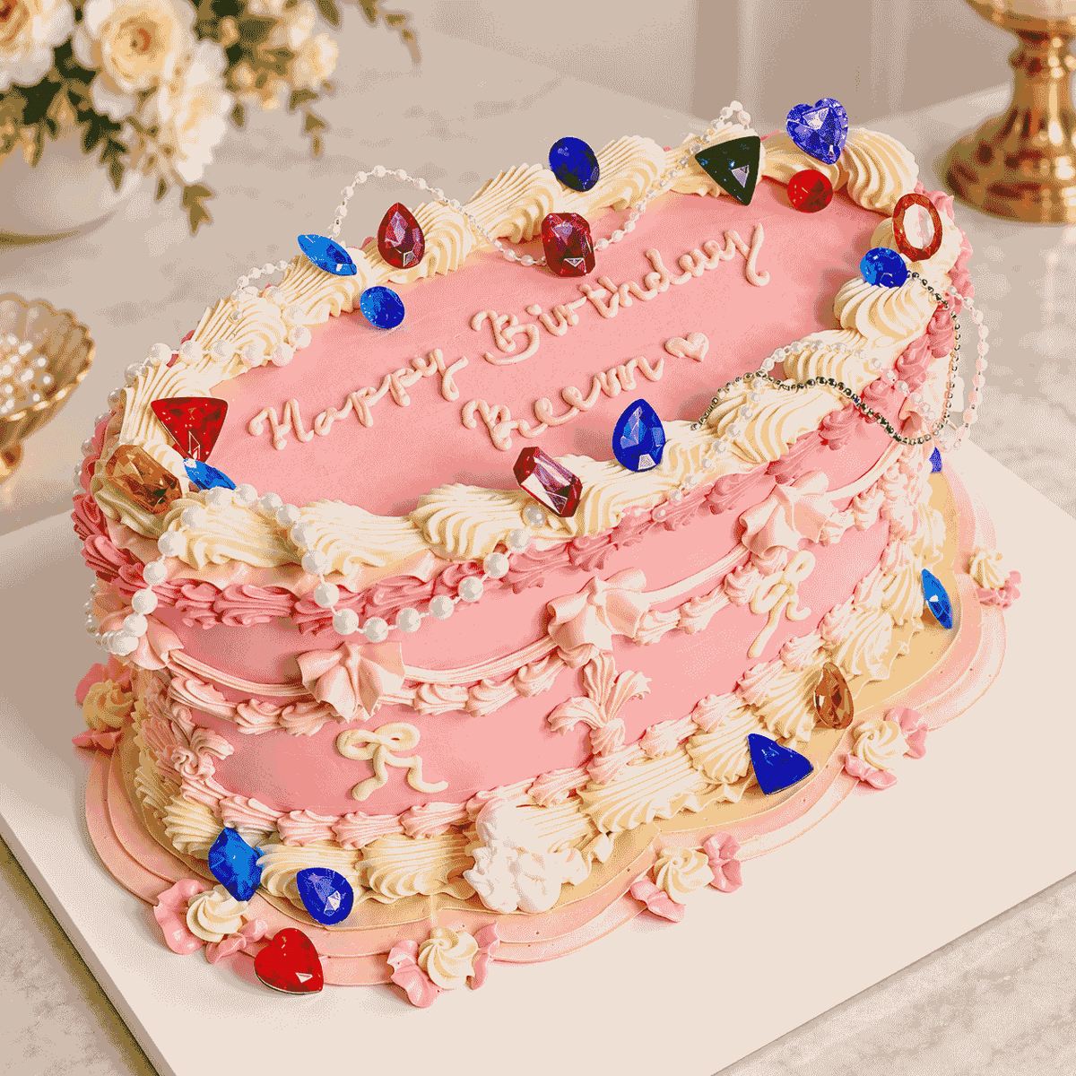 An oval pink vintage Lambeth celebration cake decorated with intricate cream piping, draped pearl bead garlands, and colorful faceted jewel stones with piped Happy Birthday script.