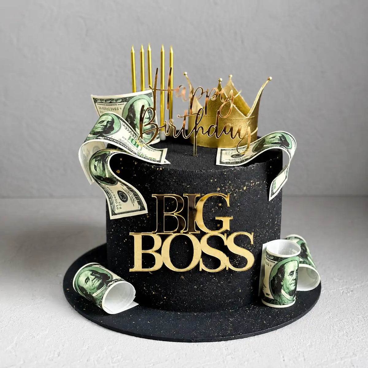 A tall round black velvet-textured birthday cake with gold splatters, a gold mirror plaque reading BIG BOSS, decorated with edible hundred-dollar bills, a gold crown, and gold candles as seen in image.