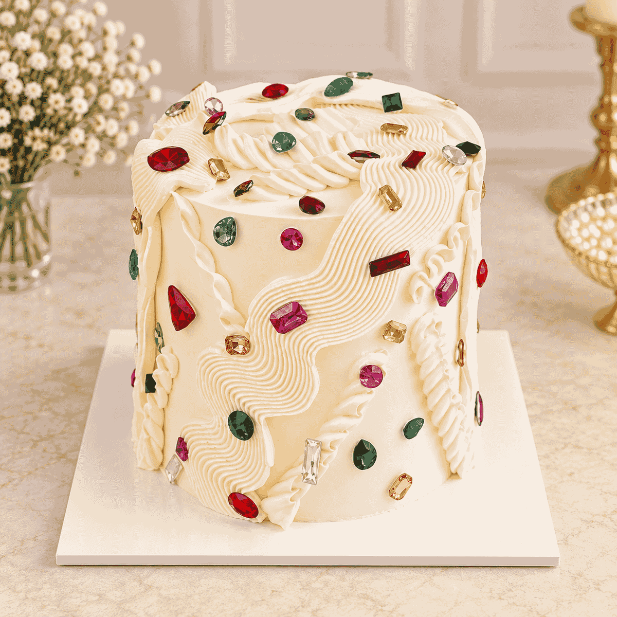 A tall ivory frosted celebration cake featuring fluid hand-piped textured ribbon waves embellished with colorful sparkling faceted jewel stones.