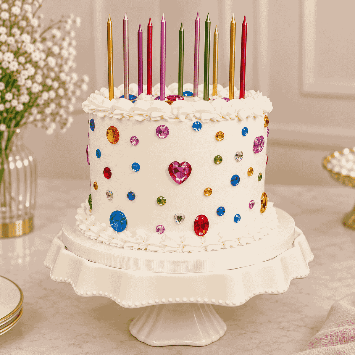 A tall ivory round celebration cake decorated with scattered colorful crystal gems, piped ruffle borders, and a row of tall slender metallic candles.
