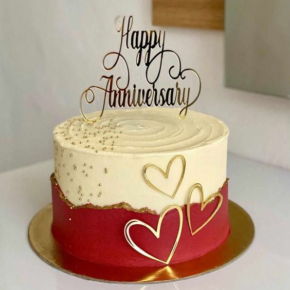 An elegant round anniversary cake with a smooth white top half and a deep red fault line bottom half trimmed with gold paint, decorated with gold micro-pearls and three gold outline hearts.