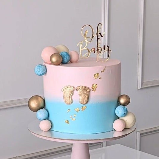 A tall pink and blue ombré baby shower cake decorated with edible gold leaf, golden baby footprints, pastel chocolate spheres, and a shiny gold 'Oh Baby' topper.