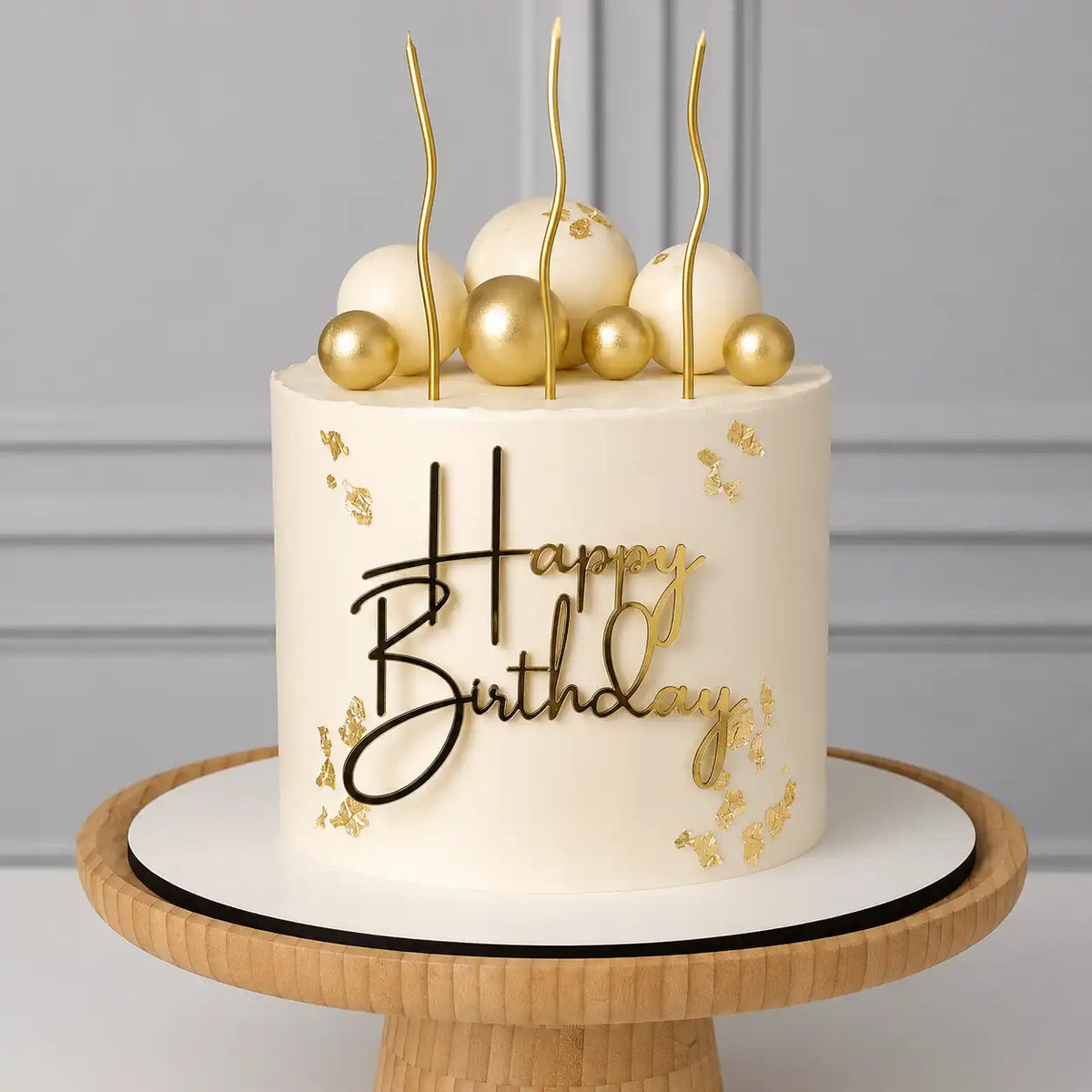 A tall round premium birthday cake with a smooth white ivory frosting body decorated with delicate gold leaf flakes, topped with white and gold spheres, a black and gold Happy Birthday topper, and three wavy gold candles as seen in image.