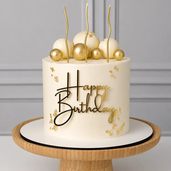 A tall round premium birthday cake with a smooth white ivory frosting body decorated with delicate gold leaf flakes, topped with white and gold spheres, a black and gold Happy Birthday topper, and three wavy gold candles as seen in image.