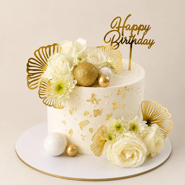 A tall round premium white birthday cake decorated with fresh white roses, chrysanthemums, scattered gold leaf flakes, metallic gold fan leaves, and polished gold spheres as seen in image.