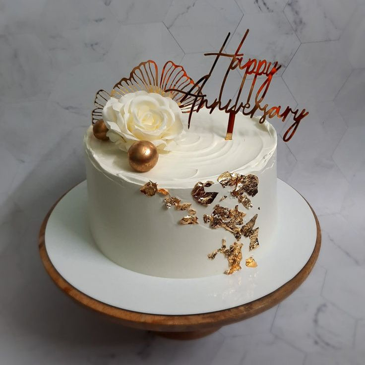 A premium white anniversary cake decorated with textured top swirls, a cluster of edible gold leaf on the side, a large white rose, metallic bronze spheres, and a gold Happy Anniversary topper.