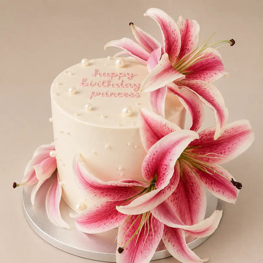 A tall round white birthday cake on a silver stand, decorated with scattered white pearls and a large, dense cascade of pink and white tiger lilies flowing down the top and sides, featuring pink script reading Happy Birthday Princess.