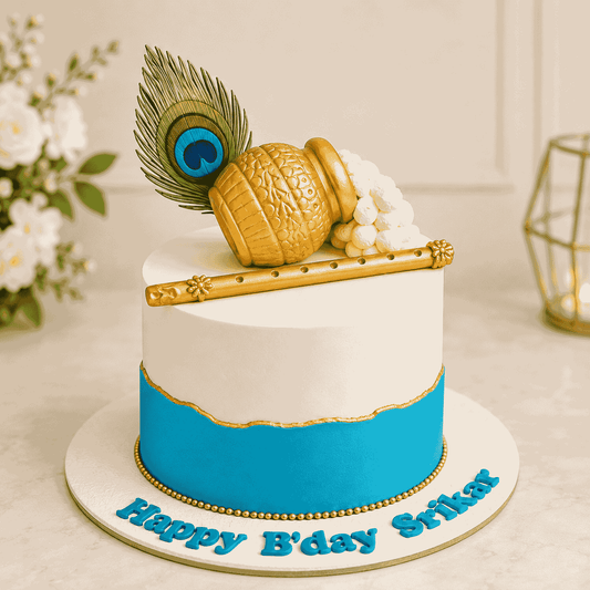 A round two-tone blue and white festive cake topped with a sculpted golden butter pot spilling white makhan, a golden flute, and a detailed peacock feather.